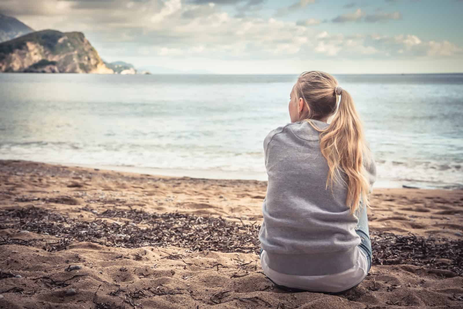 uma mulher com o cabelo comprido e loiro preso num rabo-de-cavalo senta-se na praia e olha para o mar