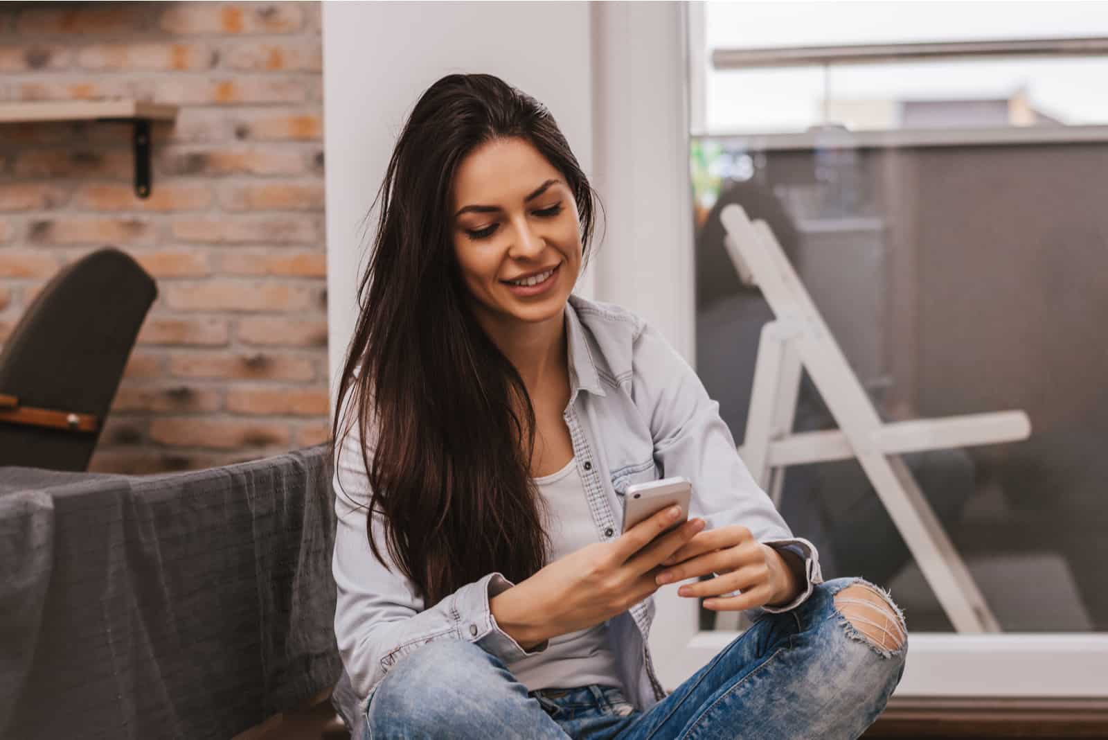 a woman with long brown hair is sitting on the couch holding a cell phone in her hand