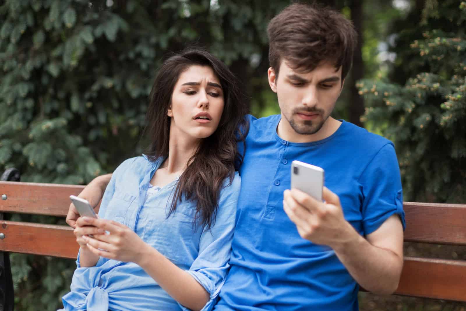 suspicious woman looking at man's mobile phone while sitting on the bench