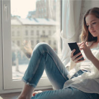 young woman sitting by the window and typing on phone