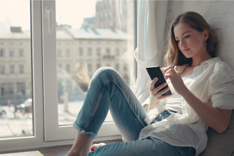 young woman sitting by the window and typing on phone