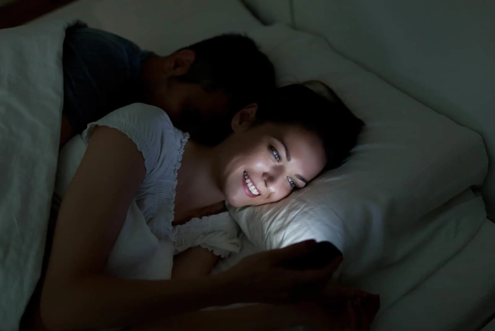 woman on phone in bed while husband is sleeping behind her