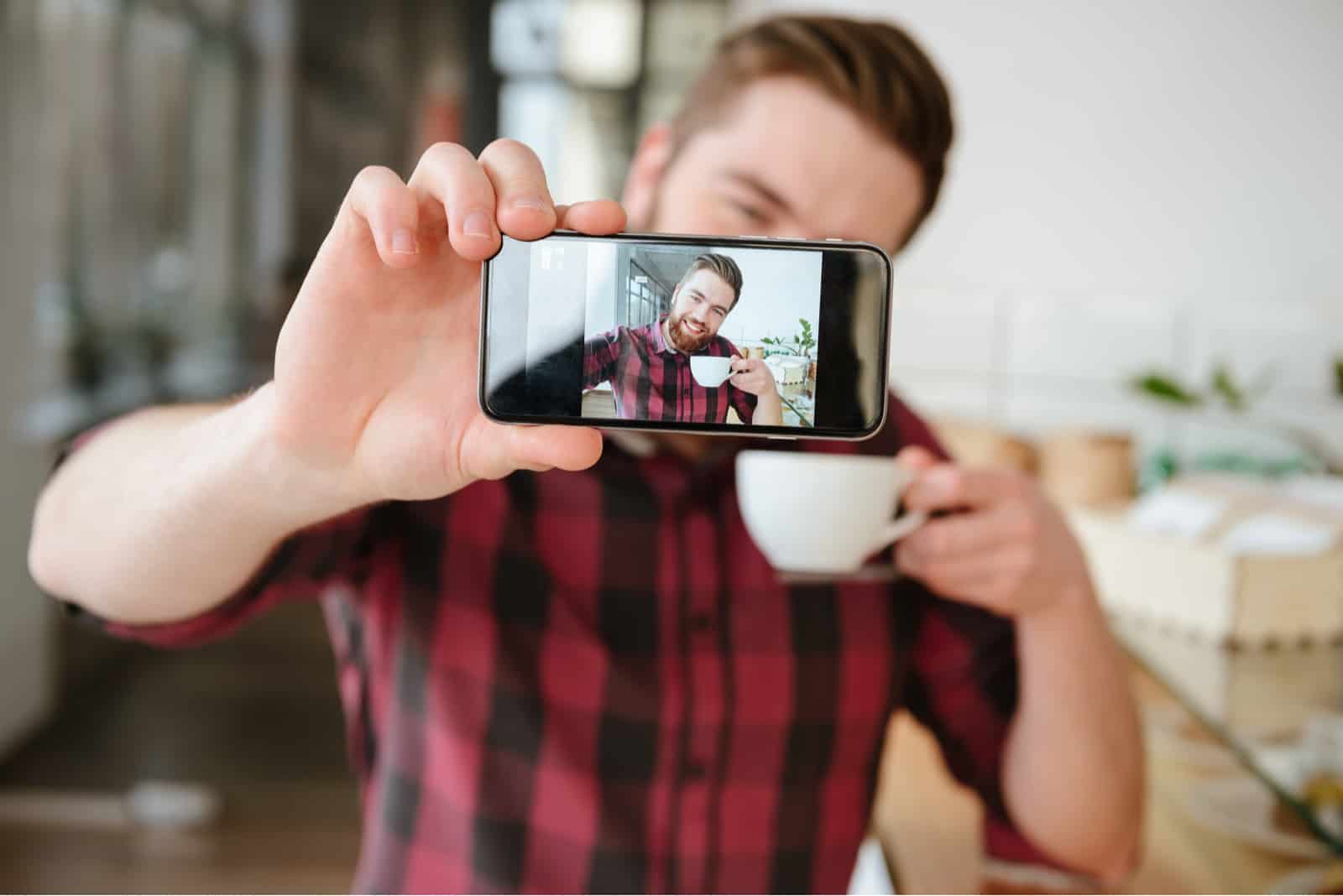 Smiling young man taking selfie and holding cup of coffee