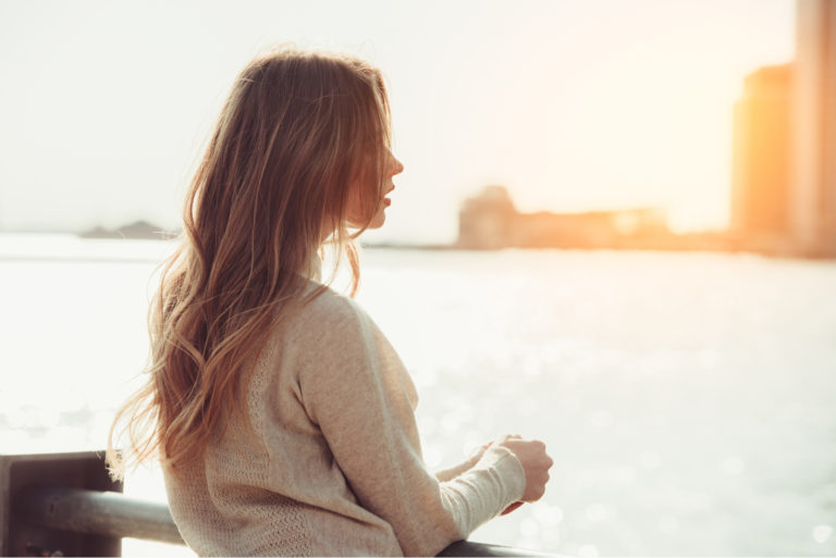 a woman with long brown hair leaning against a fence looks into the distance