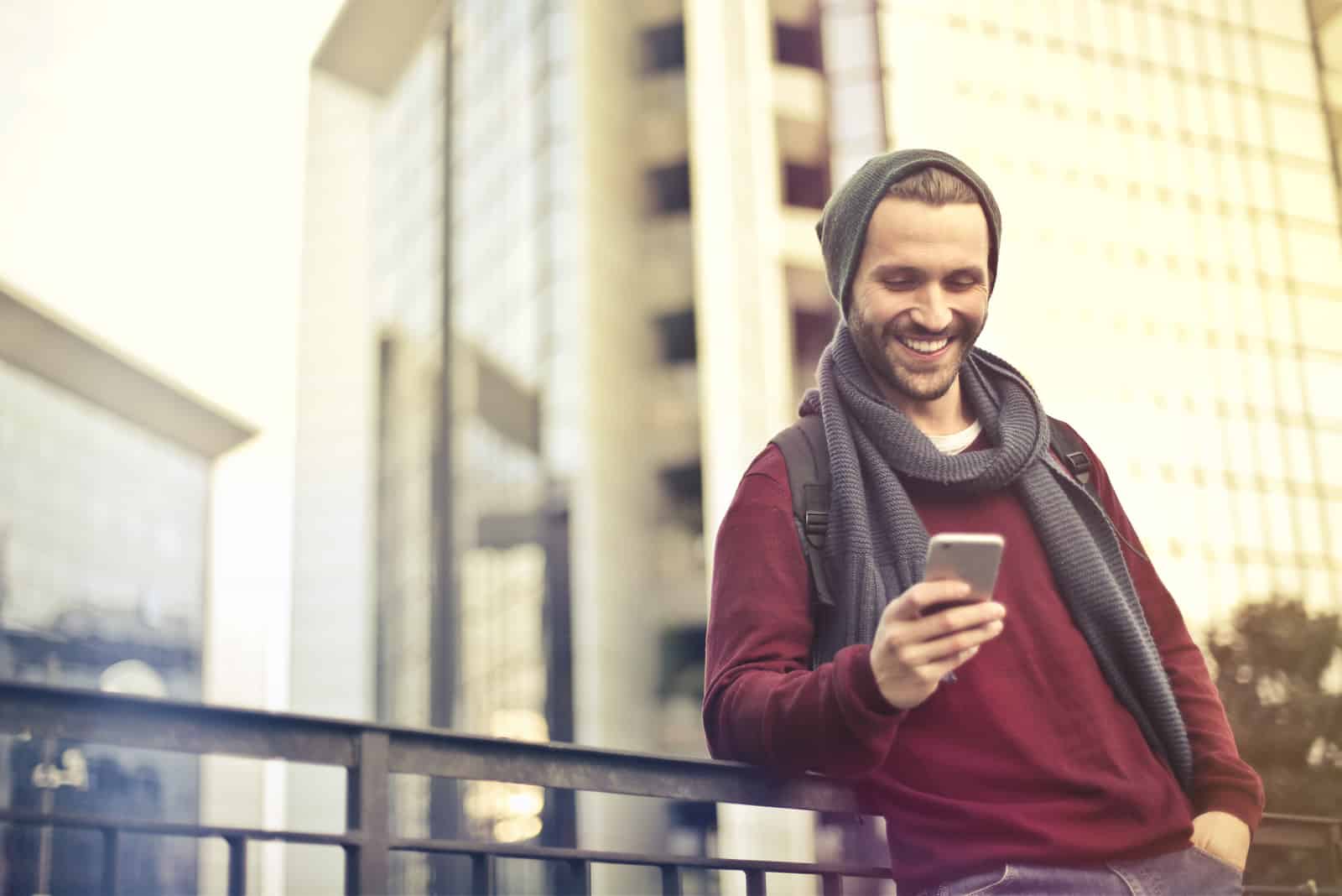 a smiling man leaning against a fence holds a phone in his hand