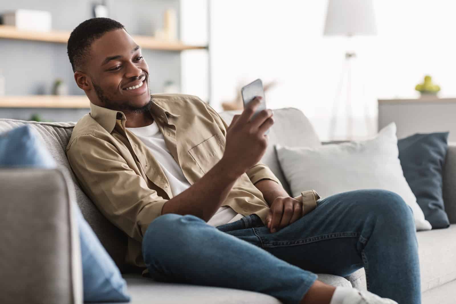 a smiling man lies on the couch and keys on the phone