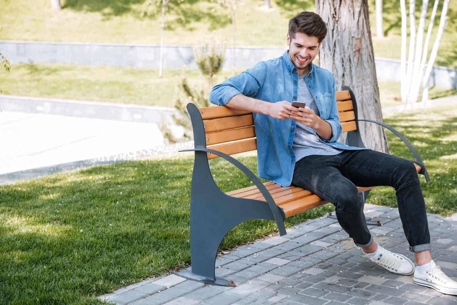 a smiling man sits on a park bench and buttons on the phone