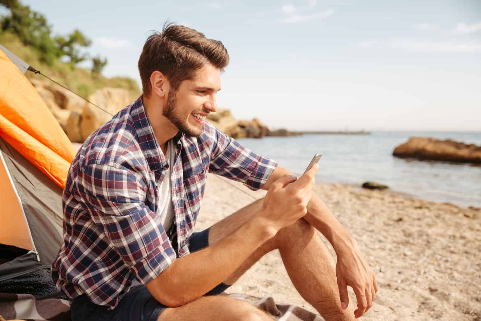 a smiling man sitting on the beach and typing on the phone