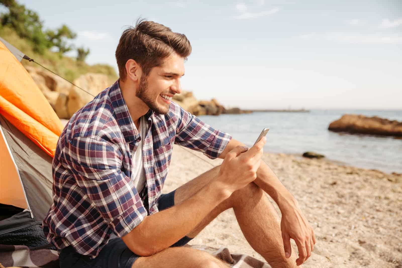 un hombre sonriente sentado en la playa y tecleando en el teléfono