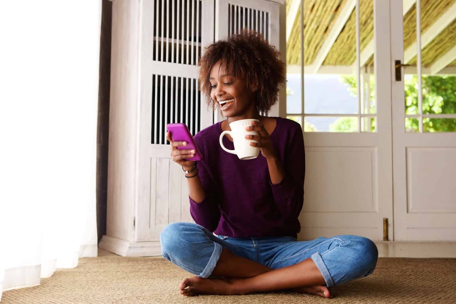 una donna sorridente seduta a terra che beve un caffè e scrive al telefono