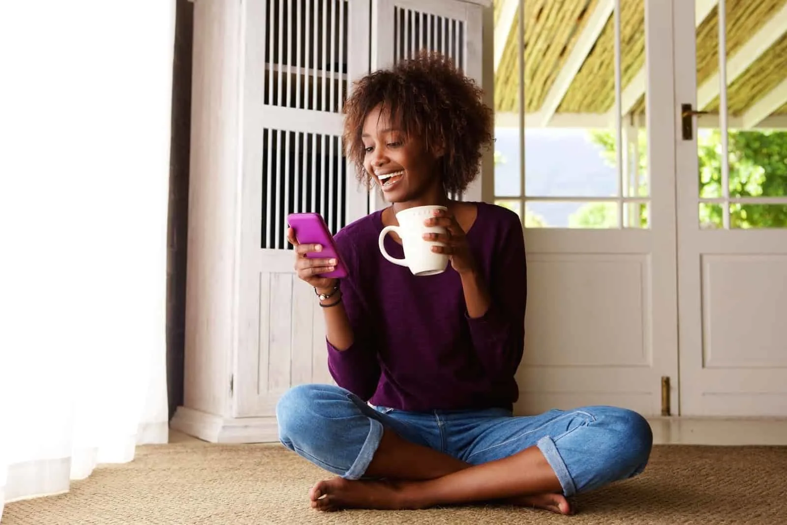 a smiling woman sitting on the floor drinking coffee and typing on the phone