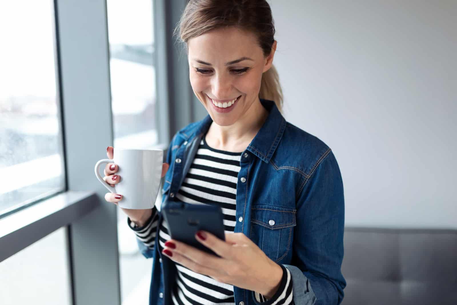 a smiling woman stands by the window and buttons on the phone
