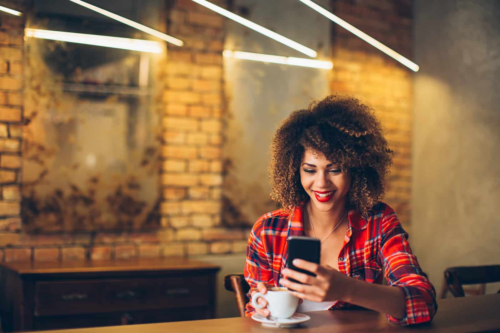 una donna sorridente con i capelli crespi seduta in un caffè a scrivere al telefono