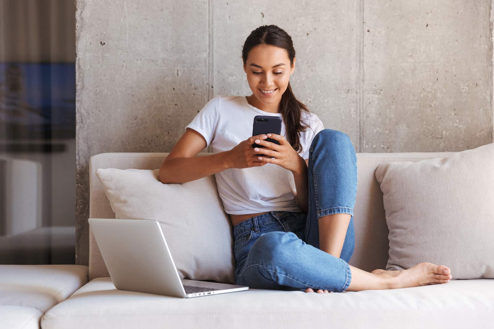 a smiling woman with tied hair is sitting on the couch holding a phone in her hand
