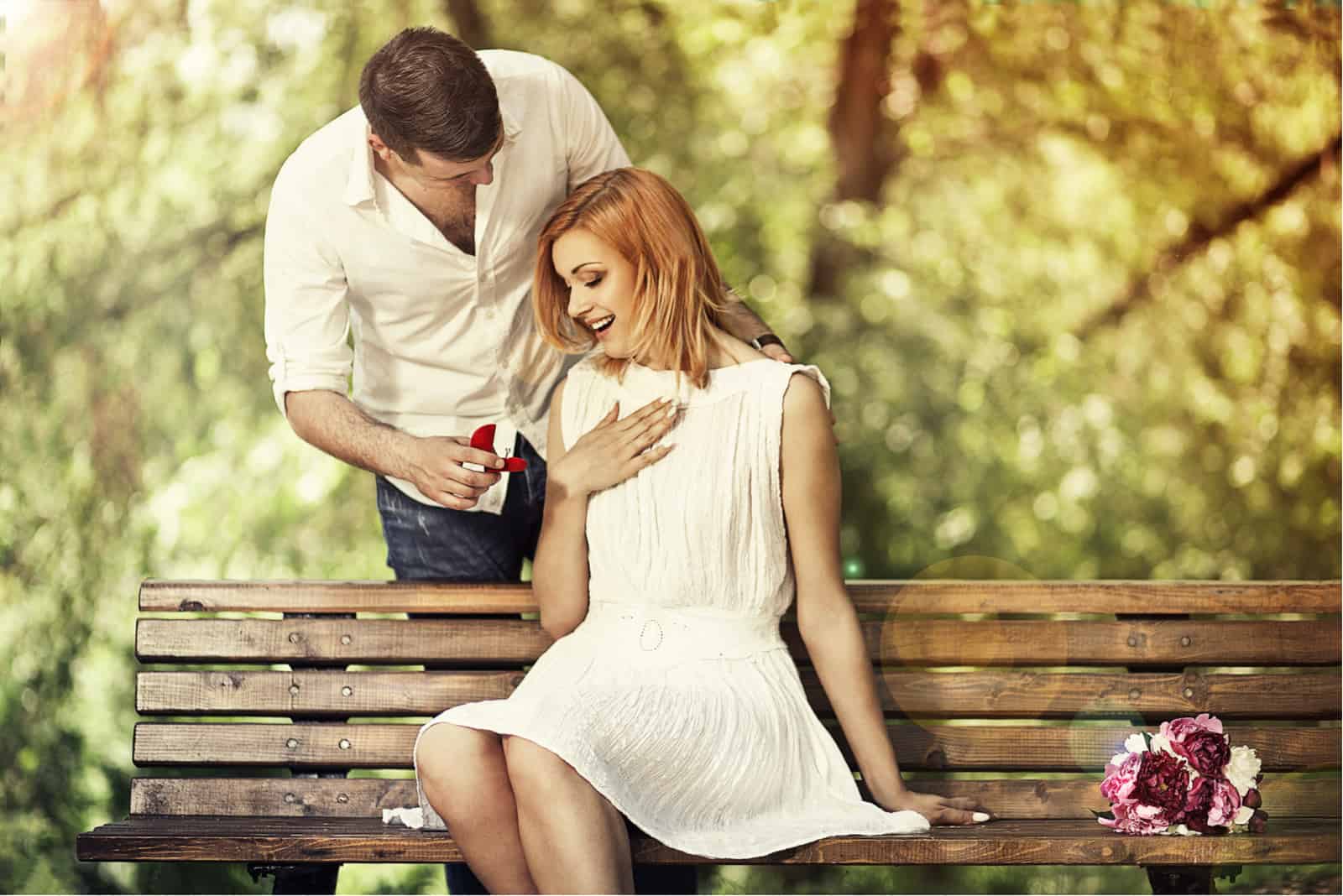 a surprised woman sits in a chair while a man gives her a ring