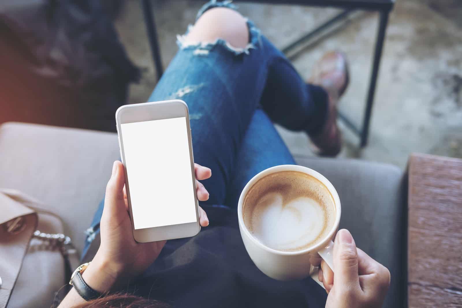 a woman sitting on the couch holding a phone and a cup of coffee in her hand