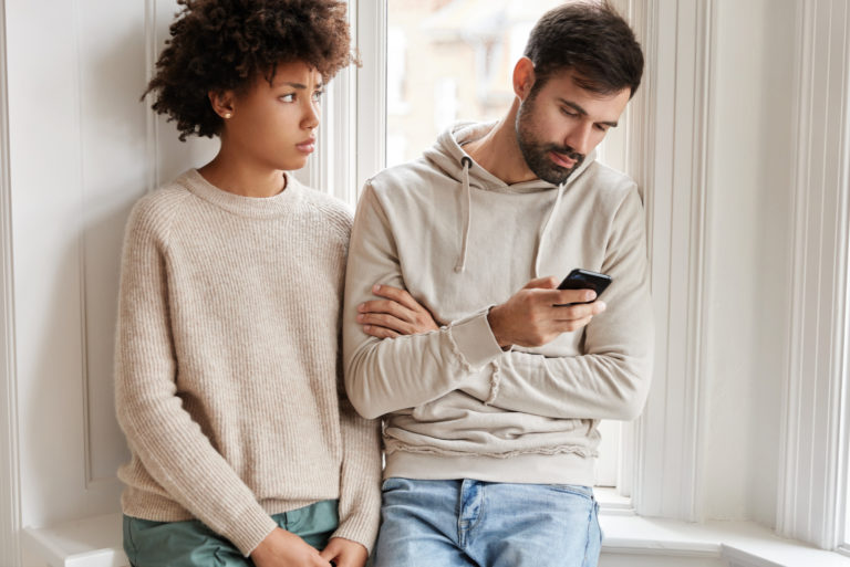 the woman stands next to the man while he ignores her and keys on the phone