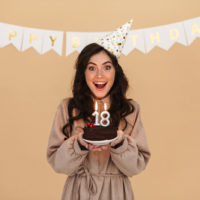 excited young woman in party cone smiling while posing with birthday cake