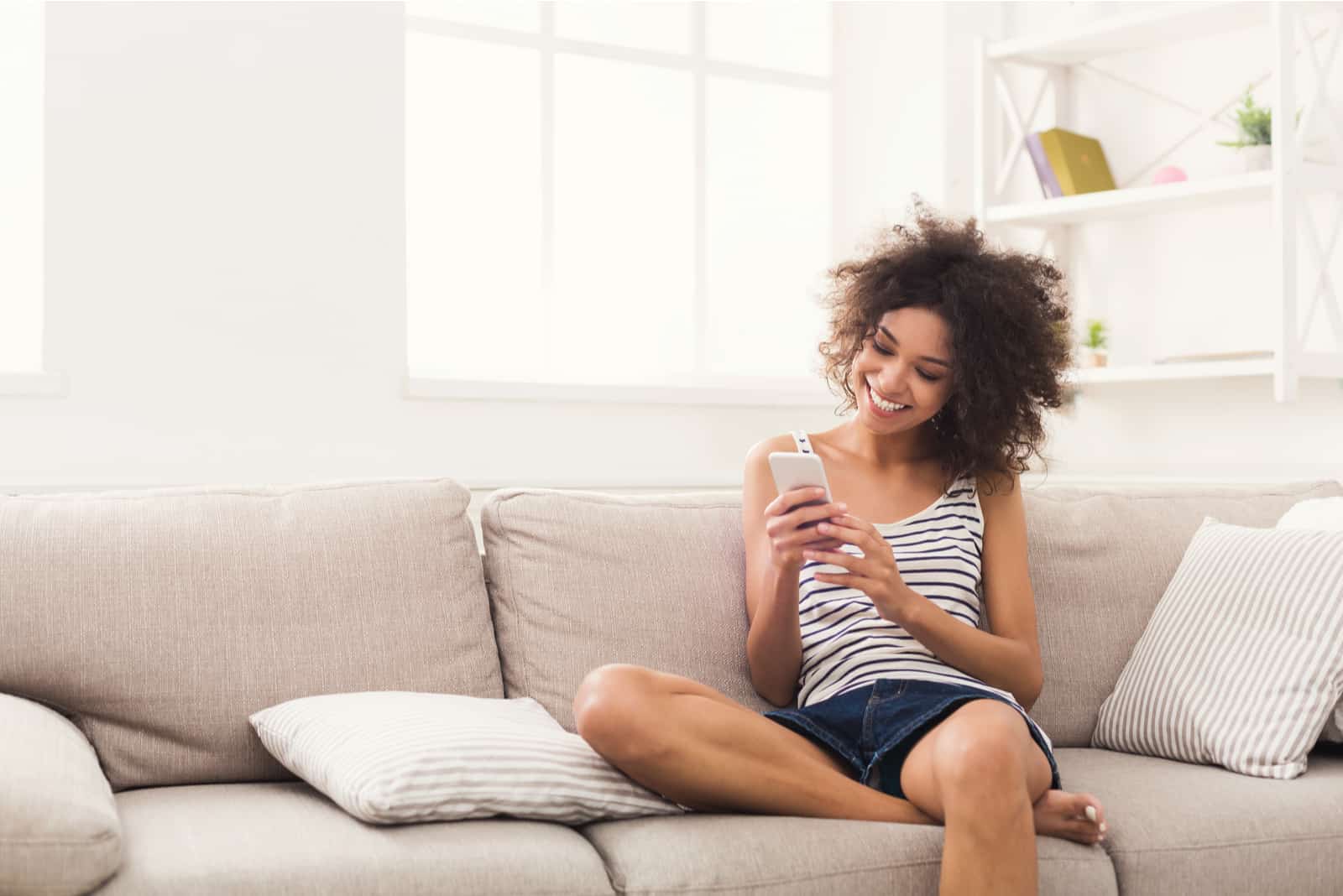 una donna sorridente con i capelli crespi si siede sul divano e attacca bottone al telefono