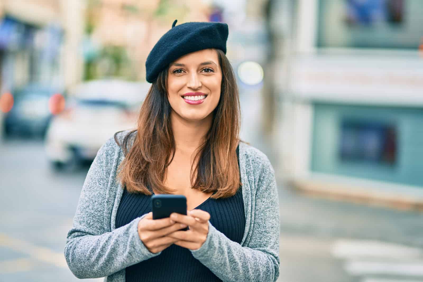 una donna sorridente con lunghi capelli castani si trova in strada con un telefono in mano