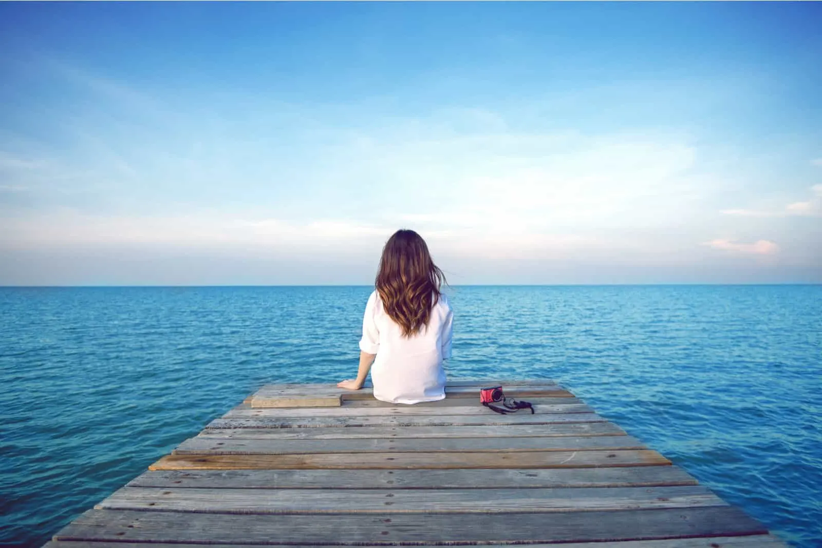 a woman with long brown hair sits on the pier and looks out to sea