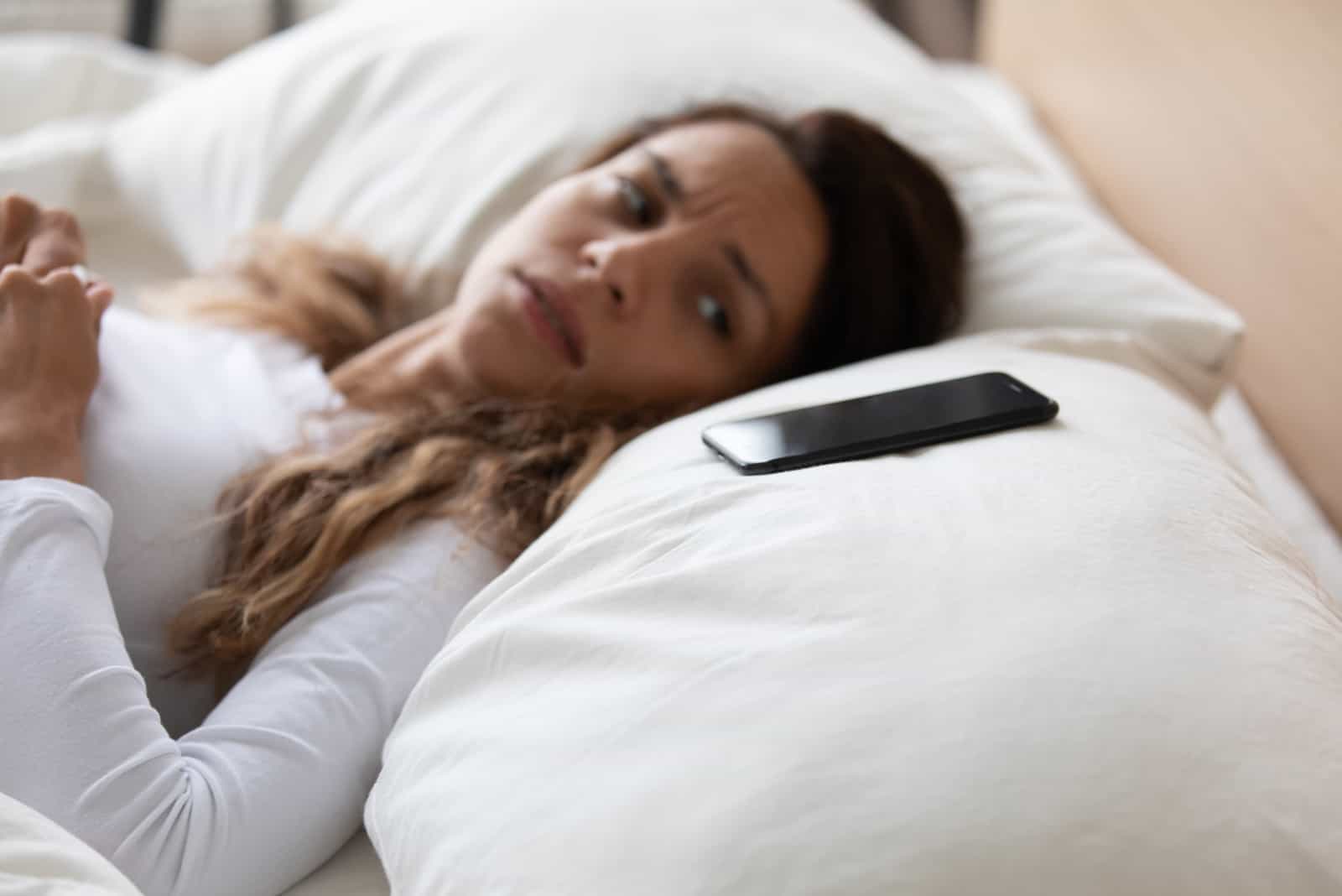 tired woman lying in bed looking at her phone on pillow