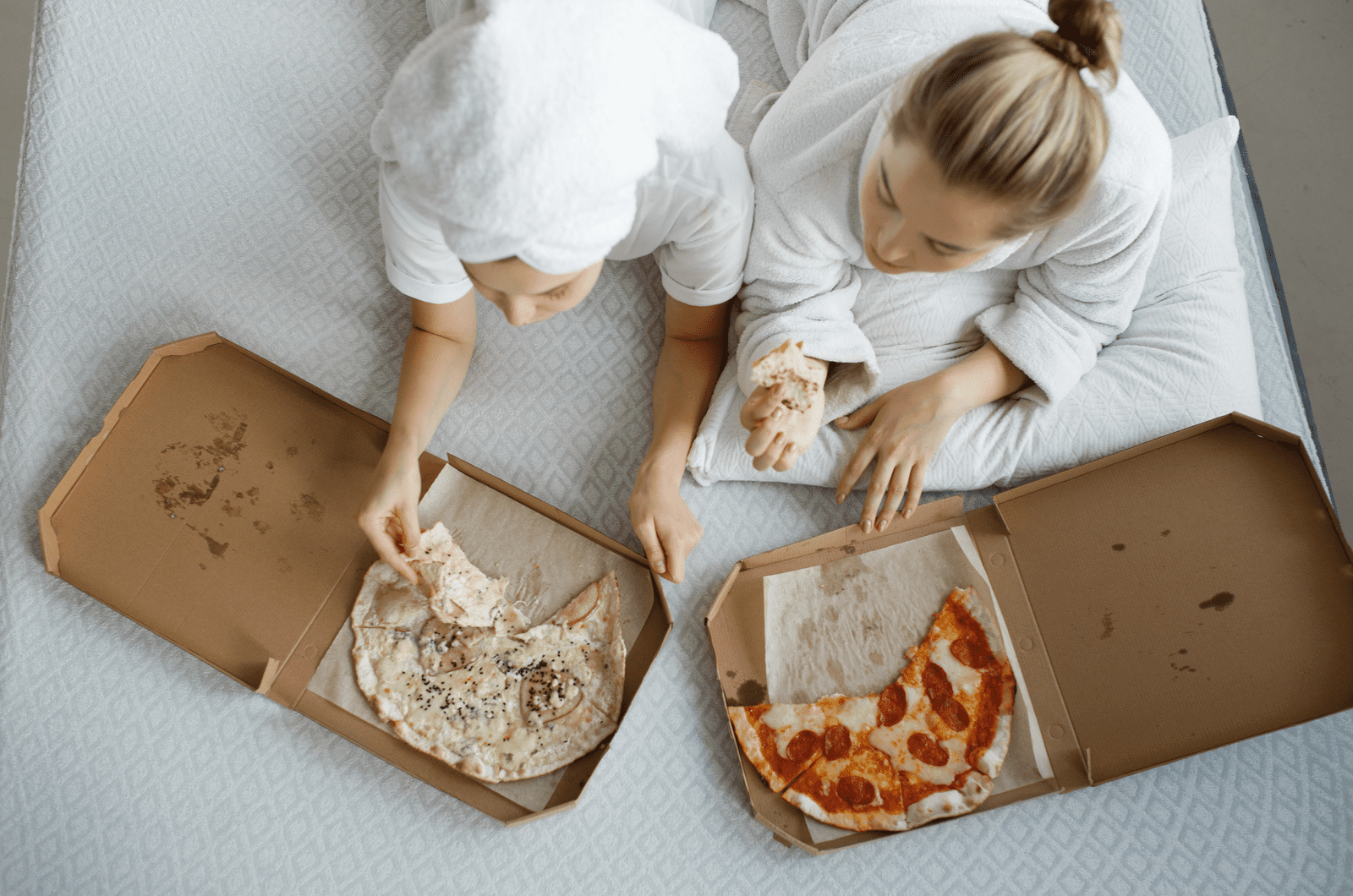 two girls in bathrobes lie on bed eating pizza and watching TV