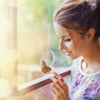 a smiling woman sits by the window and plays with a butterfly
