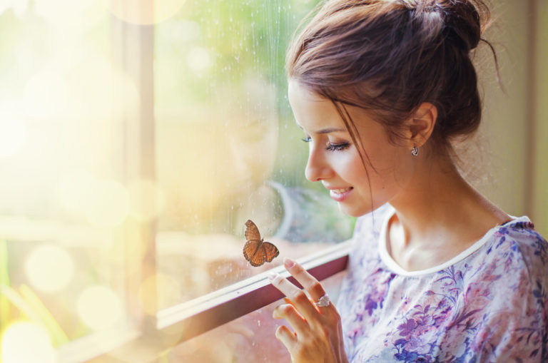a smiling woman sits by the window and plays with a butterfly