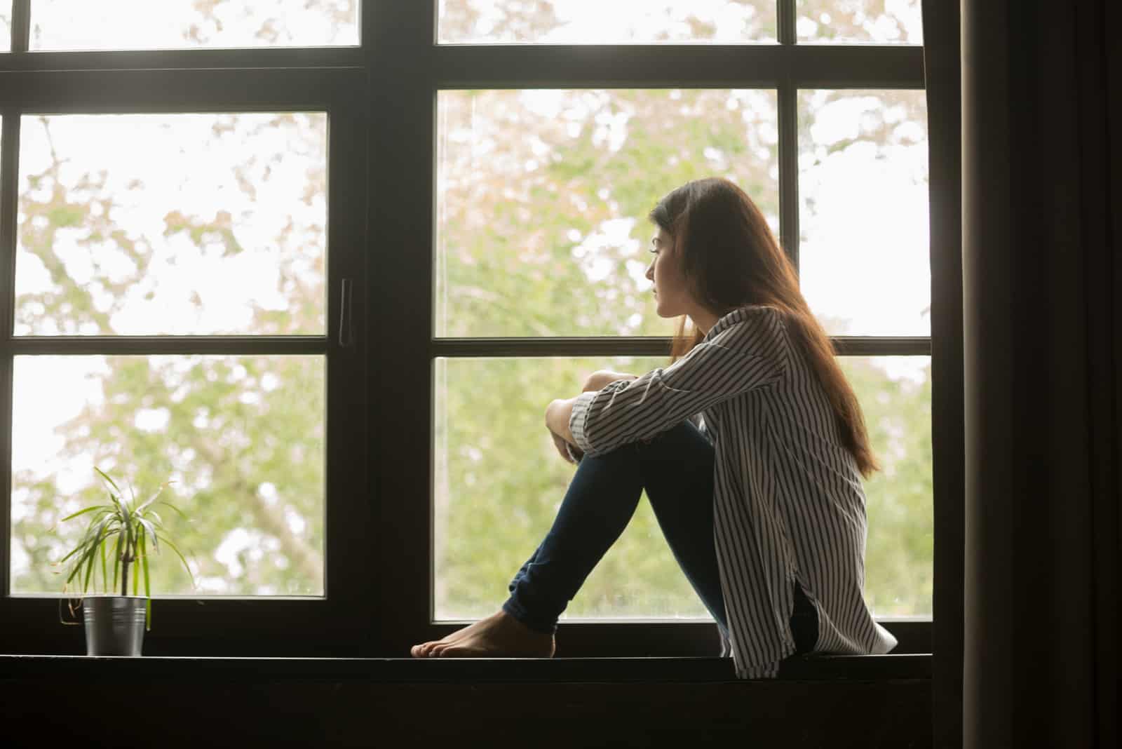 mujer joven sentada junto a la ventana sujetándose las rodillas