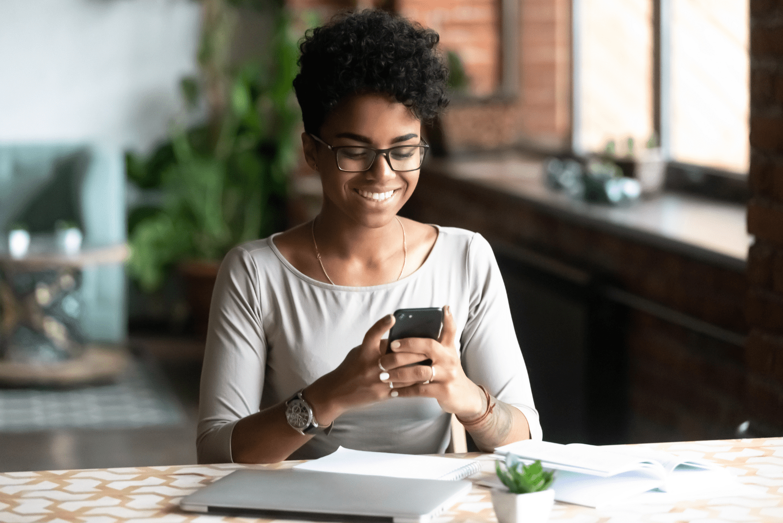 a smiling black-skinned woman sits at a table and types on a mobile phone