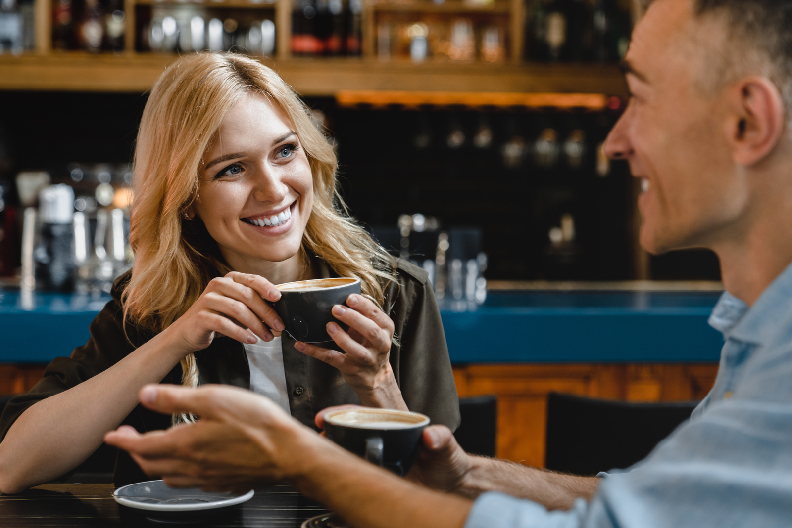 una donna sorridente con lunghi capelli biondi parla con un uomo mentre beve un caffè