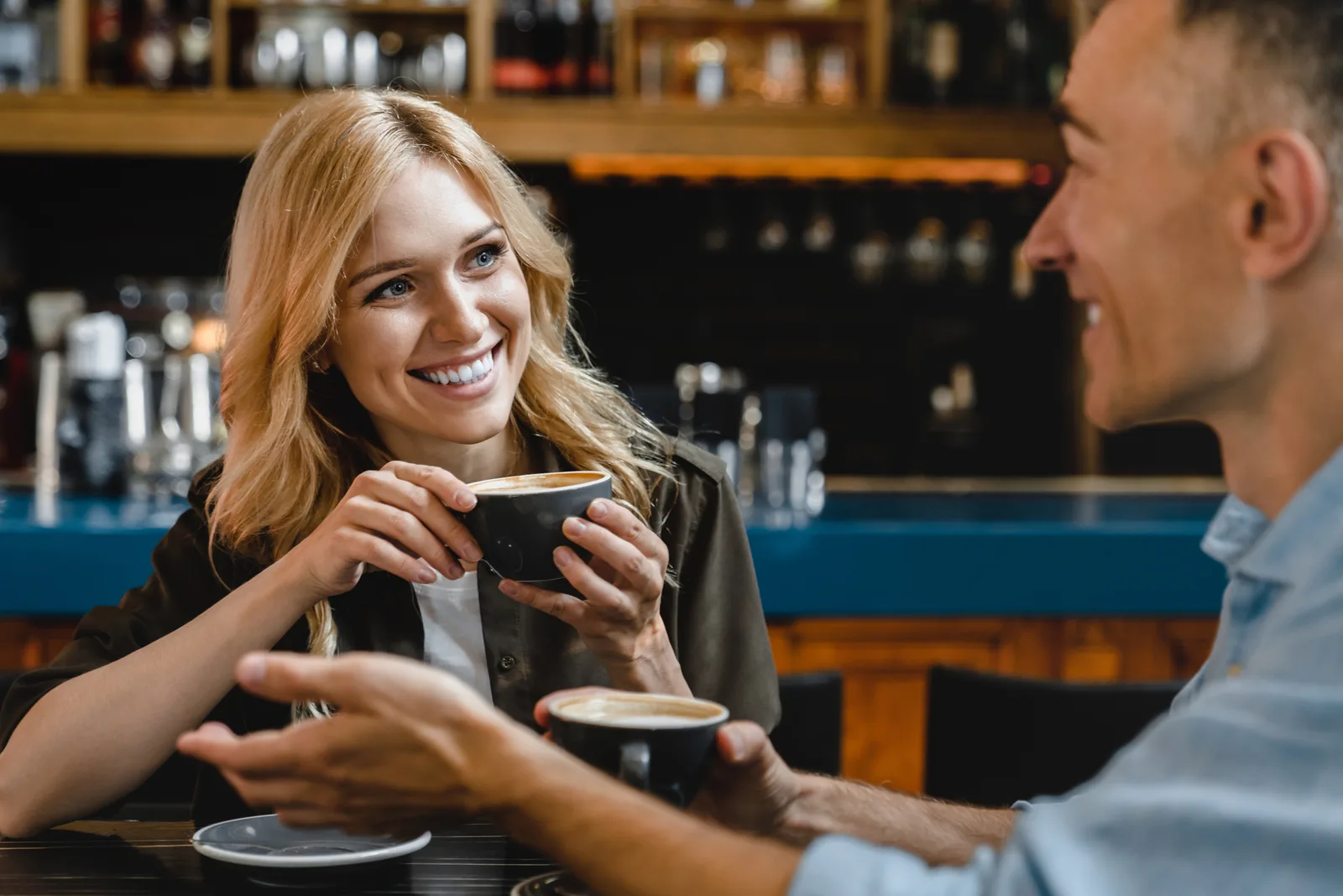 a smiling woman with long blonde hair is talking to a man while drinking coffee