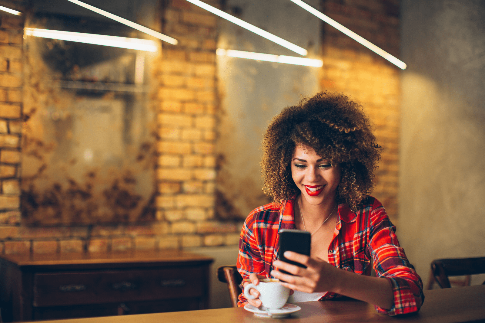 una donna sorridente con i capelli crespi è seduta in un caffè e sta scrivendo al telefono