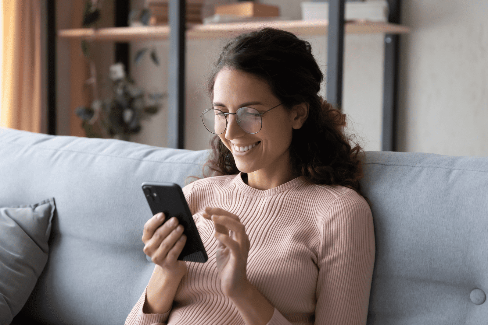 una donna sorridente con gli occhiali è seduta sul divano e digita su un telefono cellulare