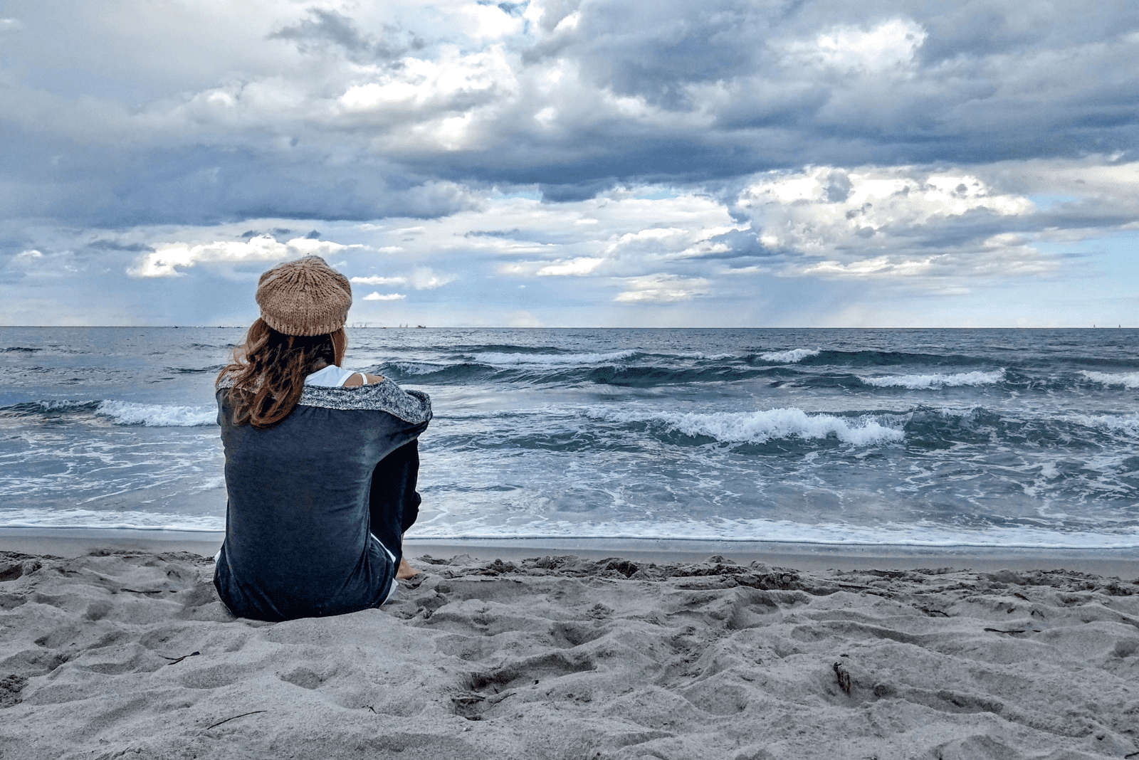 a woman with a hat on her head sits on the beach and looks at the sea