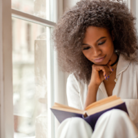 una hermosa mujer con el pelo encrespado está sentada junto a la ventana y lee un libro