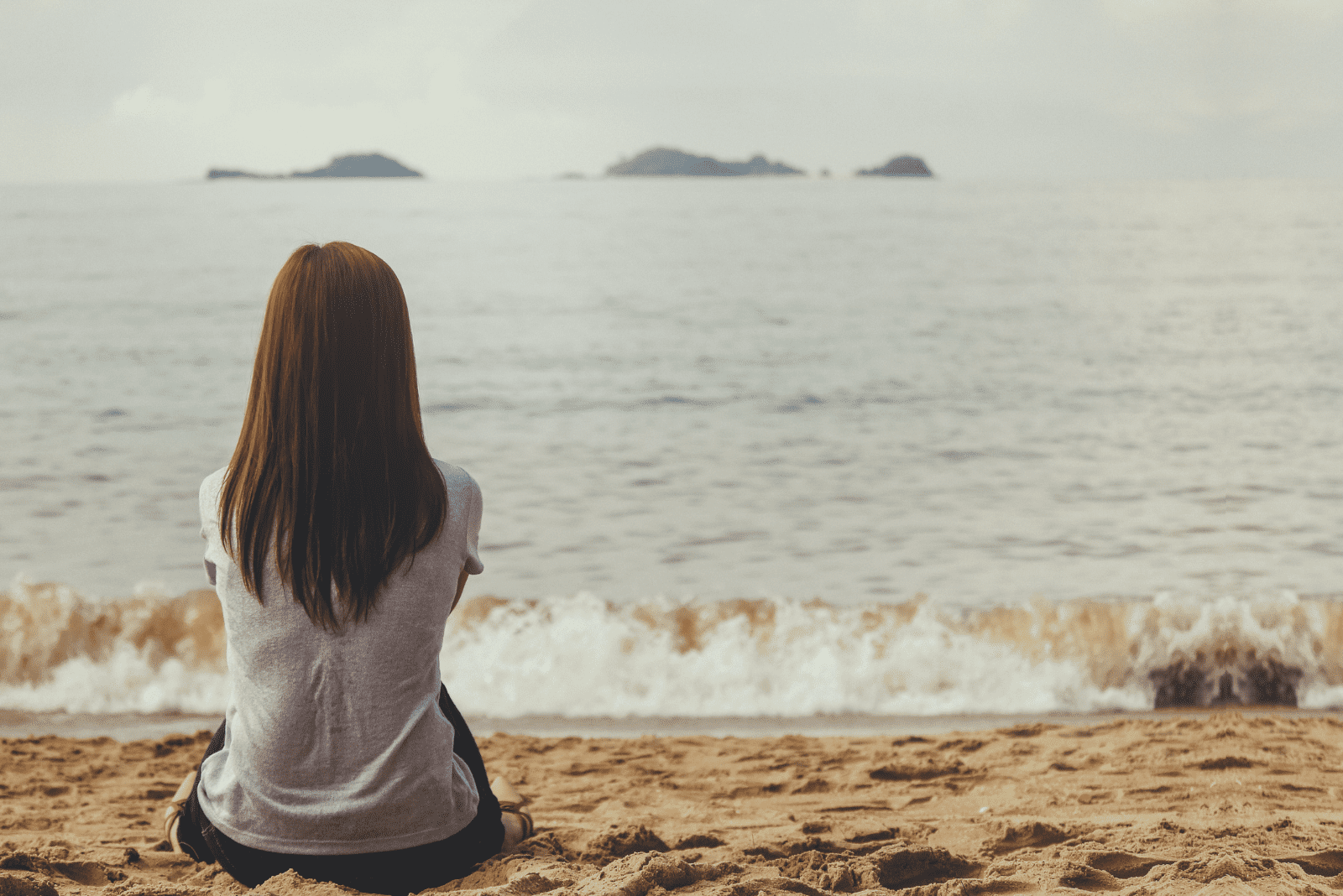 a woman with long brown hair sits on the beach and looks at the sea
