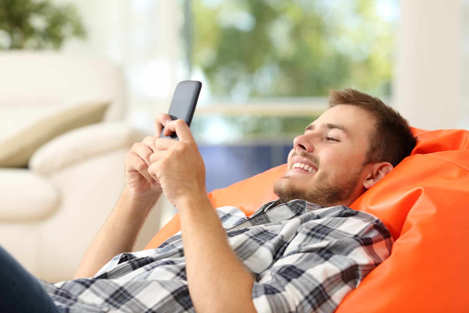 Relaxed man using a smart phone lying on an orange pouf