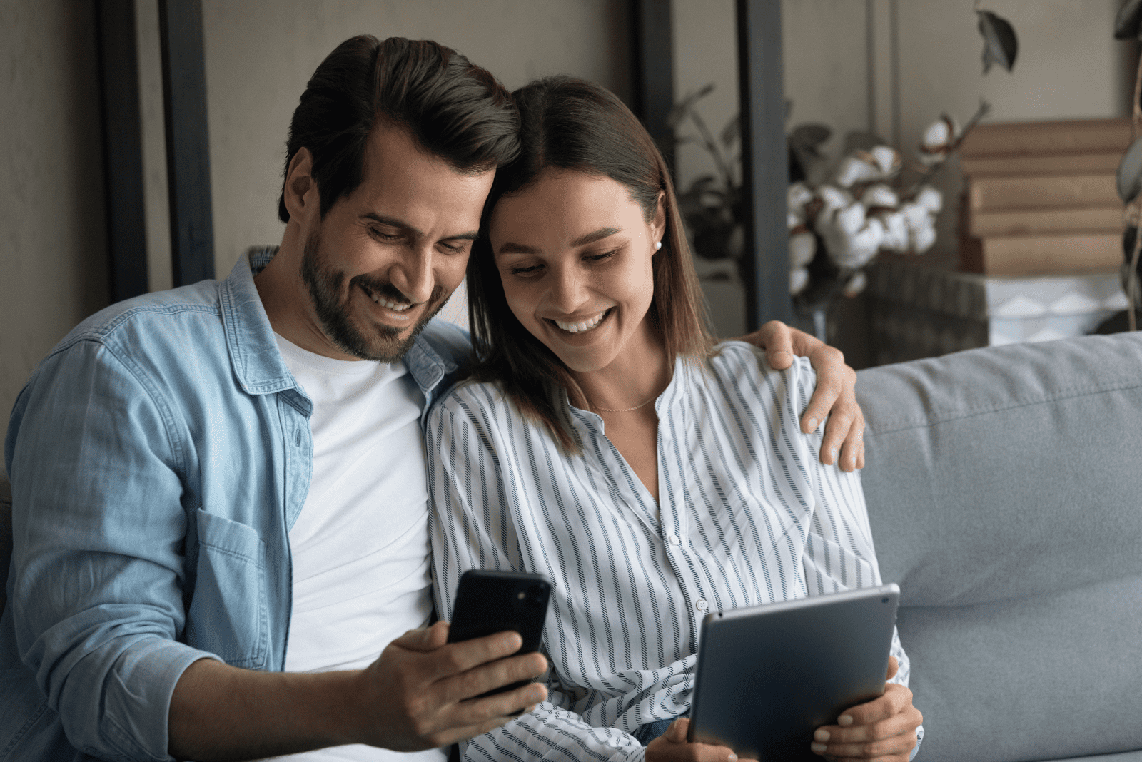 a smiling man and a woman are sitting on the couch hugging each other and looking at the phone