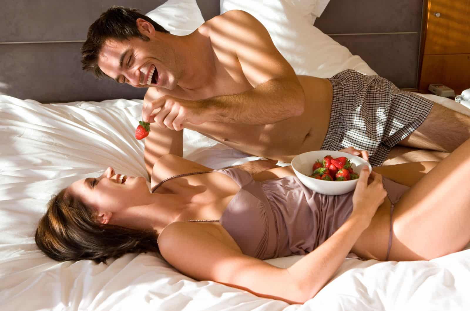 man feeding woman strawberry in bed while they're both in underwear