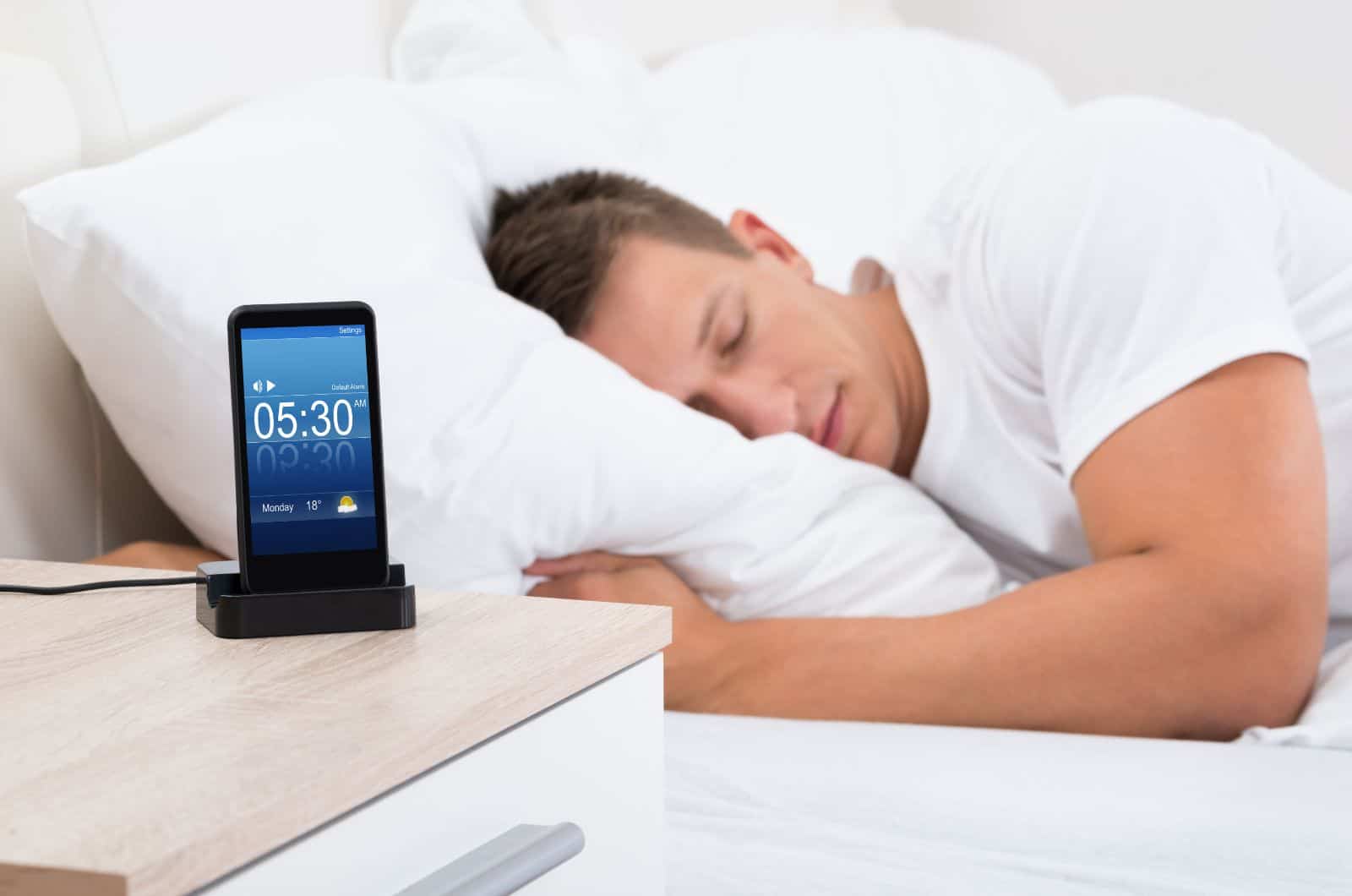 man sleeping in bed while his phone is on nightstand with alarm clock on