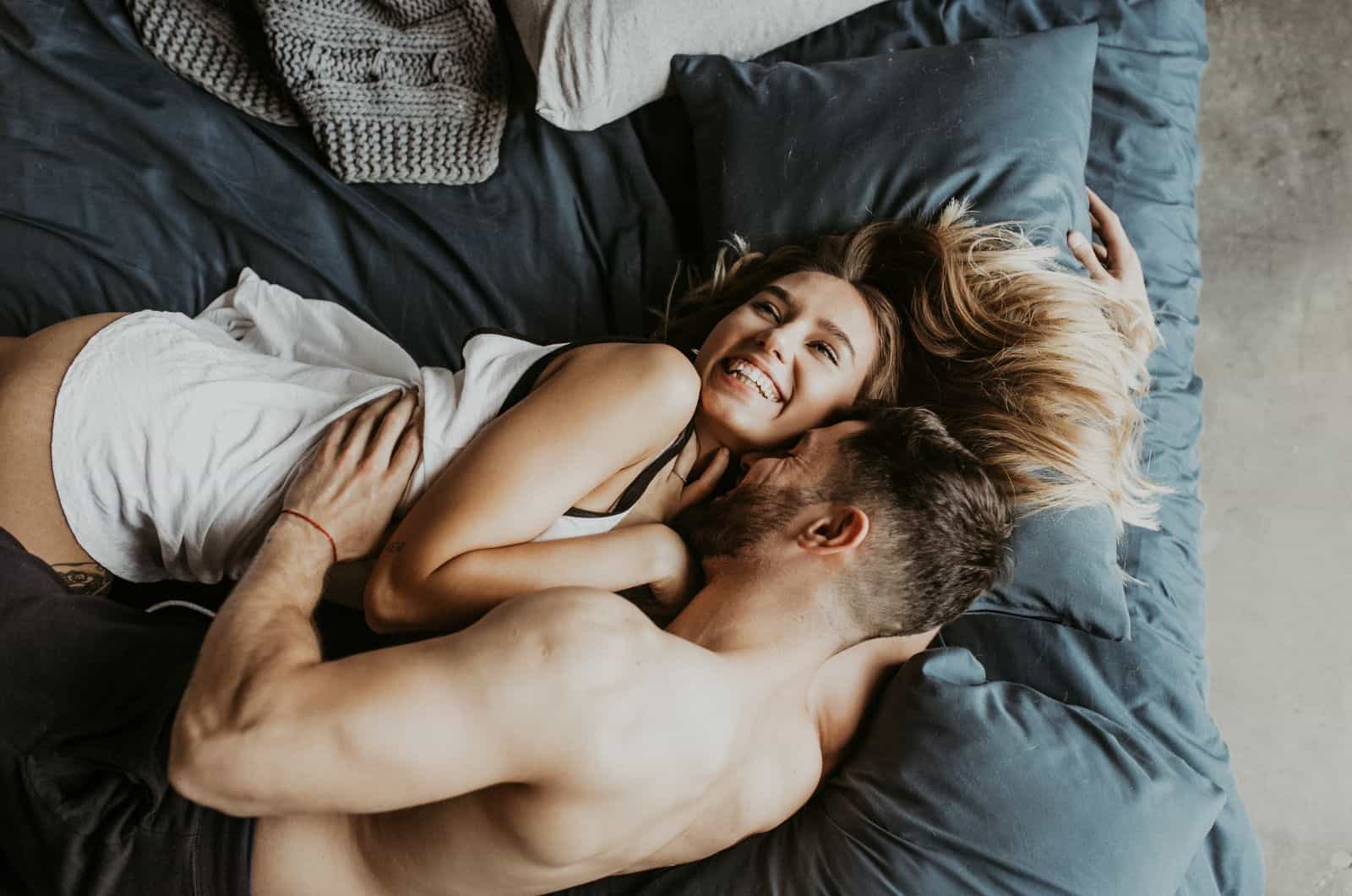 shirtless man kissing woman's neck in bed while she's smiling