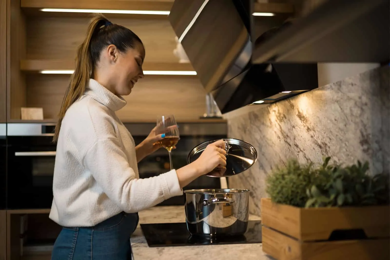 Uma bela jovem com um copo de vinho na mão olhando para a sopa que ela está cozinhando.