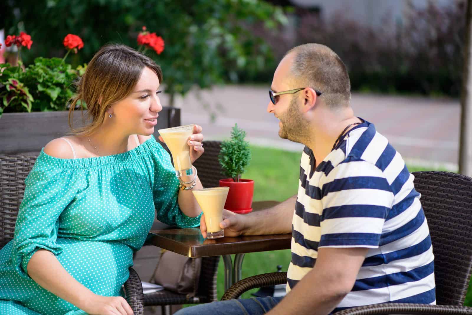 a pregnant woman sits with a man outdoors and they talk over coffee