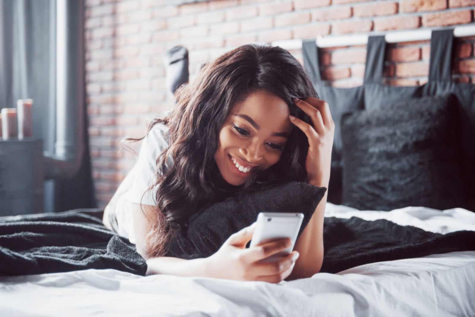 a smiling girl is lying on the bed and typing on the phone