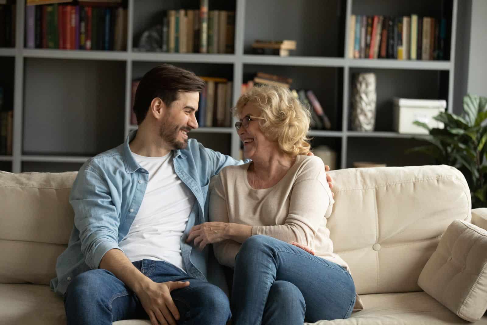 a smiling man is sitting on the couch and talking to his mom