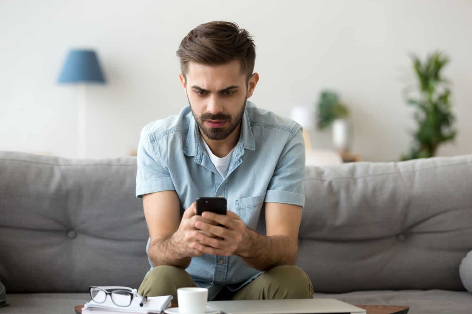 Millennial man sit on couch using smartphone shocked
