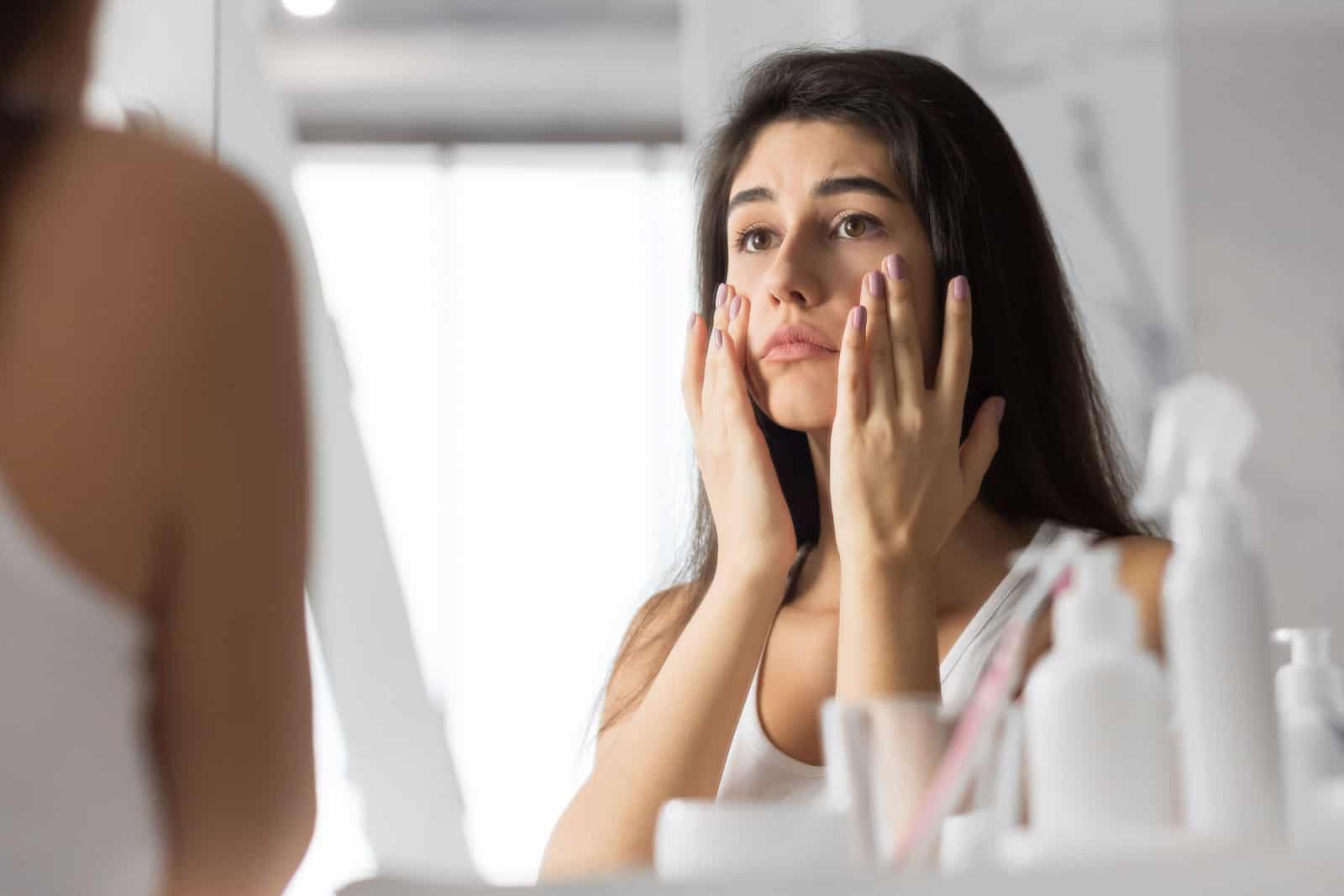 Triste giovane donna che tocca il viso guardando la sua pelle nello specchio del bagno