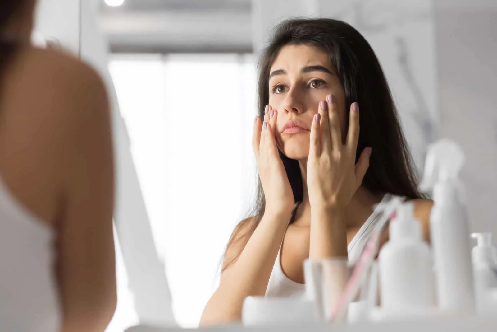 Trist ung kvinne som berører ansiktet ser på huden hennes i speilet på badet